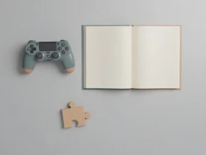 Game-controller naast een open studieboek en geometrisch puzzelstuk op een lichtgrijs bureau, bovenaanzicht.