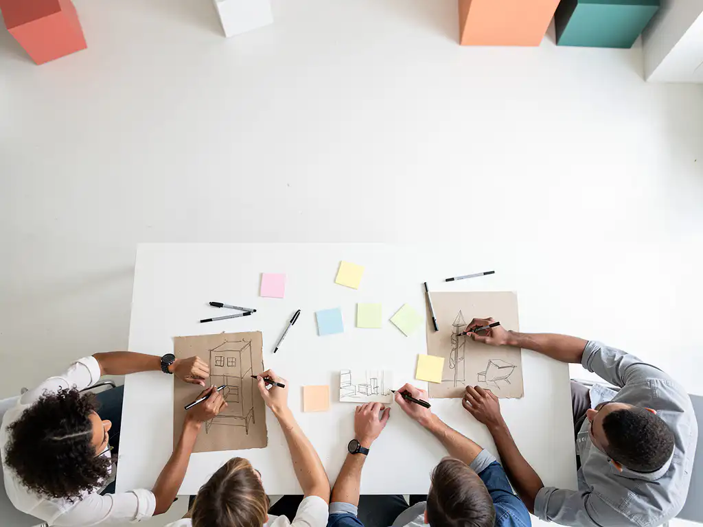 Diverse groep studenten rond een witte tafel met kleurrijke sticky notes en papieren prototypes tijdens een ontwerpworkshop.