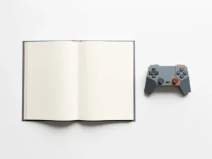 Open leerboek en geometrische gamecontroller op wit bureau, bovenaanzicht in leisteenblauw en terracotta tinten.