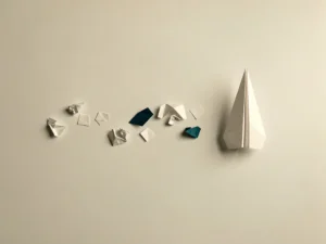 Verfrommeld papieren vliegtuigje op wit bureau naast een perfect gevouwen exemplaar, omringd door geometrische papiersnippers.