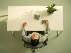 Kantoormedewerker ontspant achteroverleunend in minimalistische stoel, ogen gesloten, met gamecontroller en plant op wit bureau.