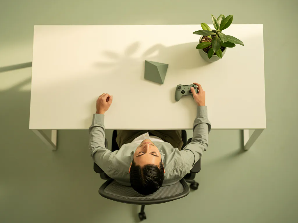Kantoormedewerker ontspant achteroverleunend in minimalistische stoel, ogen gesloten, met gamecontroller en plant op wit bureau.