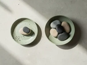 Twee minimalistische keramische kommen van bovenaf gefotografeerd, één met een enkele steen, de ander met meerdere stenen, op betonnen ondergrond.