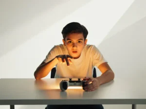 Student leunt geconcentreerd over bureau met game controller, gezicht verlicht door warm amberkleurig scherm.